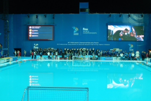 Eine Gruppe von Menschen um ein Hallenbad mit Netz am Boden herumstehend, mit einem Bildschirm im Hintergrund, der "FINA World Championships 2015 Kazan" anzeigt.