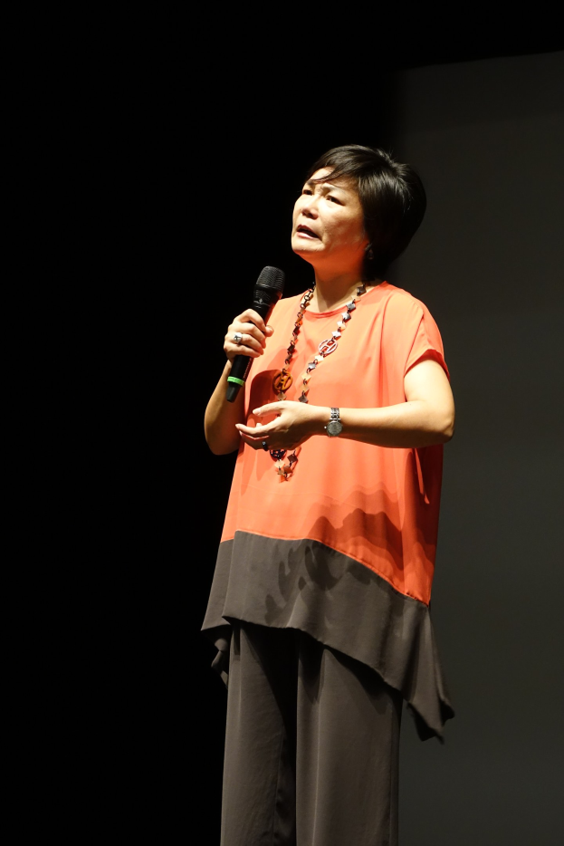 Frau auf einer hell erleuchteten Bühne mit einem Mikrofon in der Hand, die eine weiße Bluse und einen schwarzen Rock trägt und ihr Haar zu einem Knoten gebunden hat, während sie selbstbewusst in die Menge spricht.