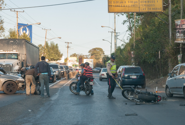 Eine Gruppe von Menschen steht um ein verunglücktes Motorrad auf der Straße herum, umgeben von mehreren Fahrzeugen, darunter ein Lastwagen, und einer Kulisse aus Bäumen, Polen, Lampen, Schildern und Himmel.