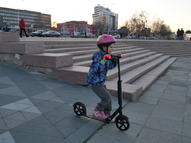 Ein junges Mödchen in einem Helm fährt mit einem Scooter eine Treppe hinunter, mit Fahrzeugen, Menschen, Laternen, Bäumen, Gebäuden und einem klaren blauen Himmel im Hintergrund.