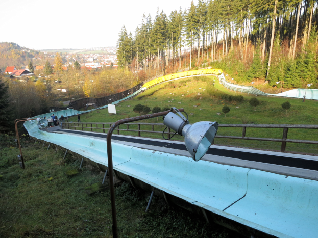 Ski-Schanze mit einer Leuchte oben drauf, Menschen, die runterrutschen, umgeben von Bäumen und Häusern auf einer grünen Wiese unter einem sichtbaren Himmel.