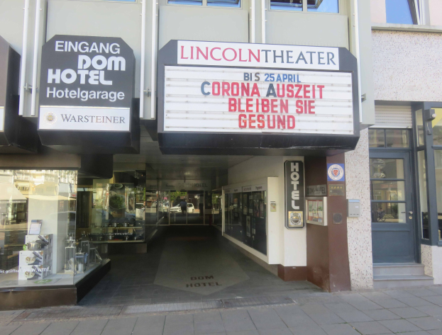 Außenansicht des Lincoln Theaters in Berlin, Deutschland, mit Glasfenstern und -türen sowie einer Tafel mit Text und einer belebten Stadtansicht im Inneren.