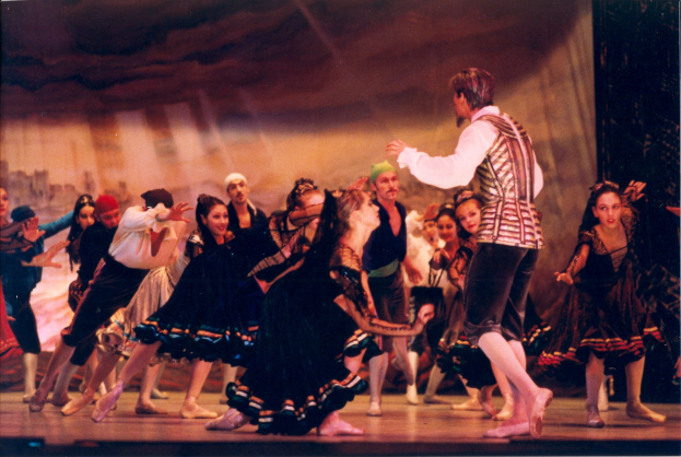 Eine Gruppe von Menschen in bunten Kostümen tanzt auf einer hell erleuchteten Bühne mit einer Wand im Hintergrund.