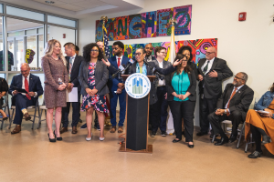 Gruppe von Menschen vor einem Rednerpult mit Mikrofon und einer Tafel mit der Aufschrift 'Sancho-Stadtrat tritt dem Stadtrat bei', mit sitzenden und stehenden Teilnehmern, Flaggen, Feuerlöscher und Deckenbeleuchtung im Hintergrund.