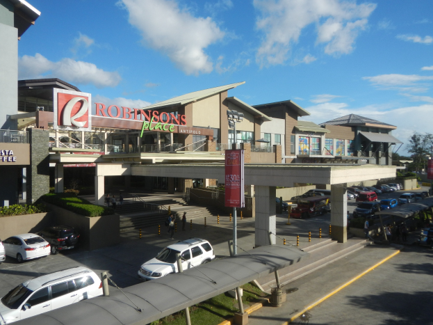 Ein belebtes Einkaufszentrum mit Gebäuden, Ladengeschäften, Fahrzeugen, Fußgängern, Straßenmöblierung, Grünflächen und einem bewölkten Himmel.