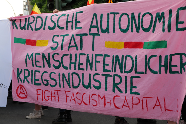 Eine Gruppe von Menschen mit einer pinken Fahne, auf der 'Kurdische Autonomie Statt Menschenfeindlicher Kriegsindustrie Fight Fascism Capital' steht, auf einer Straße mit Ampeln, Flaggen, Bäumen, Gebäuden und einem klaren blauen Himmel im Hintergrund.
