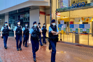 Eine Gruppe von Polizisten in Masken geht eine Straße vor einem Glasgebäude entlang, mit einem Geschäft auf der rechten Seite, das Plakate an seinen Glaswänden hat.