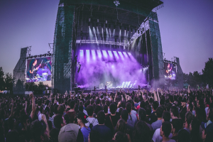 Eine große Menschenmenge auf einem Musikfestival mit einer Bühne, auf der Musiker spielen, beleuchtet von Scheinwerfern und umgeben von Bildschirmen, vor einer Kulisse aus einem Gebäude, Bäumen und einem klaren blauen Himmel.