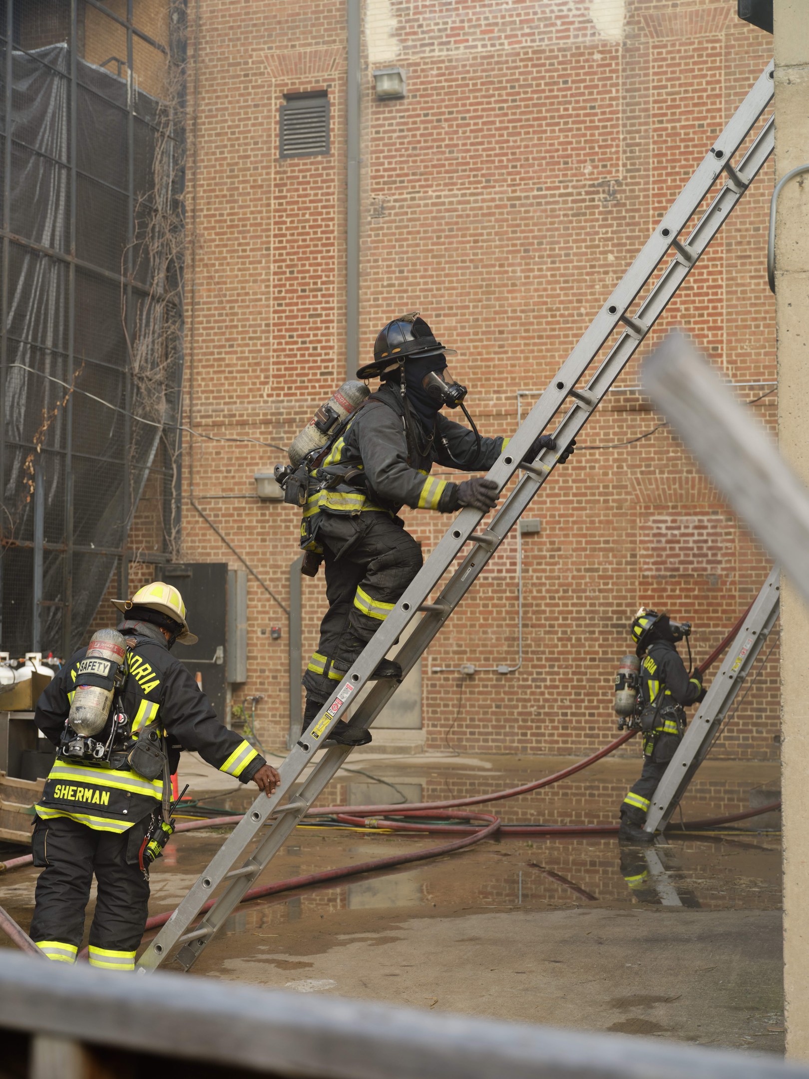 Feuerwehrleute in Helmen und Lufttanks klettern an einer Leiter vorbei an einem Backsteingebäude mit freiliegenden Rohren und einer Metallstange am Boden, während im Hintergrund ein weiteres Gebäude mit Fenstern und einem Netz zu sehen ist.