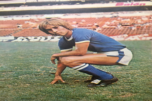 Ein Mann kniet auf einem Fuballfeld umgeben von Menschen, die auf Stühlen und Platten mit Text sitzen.