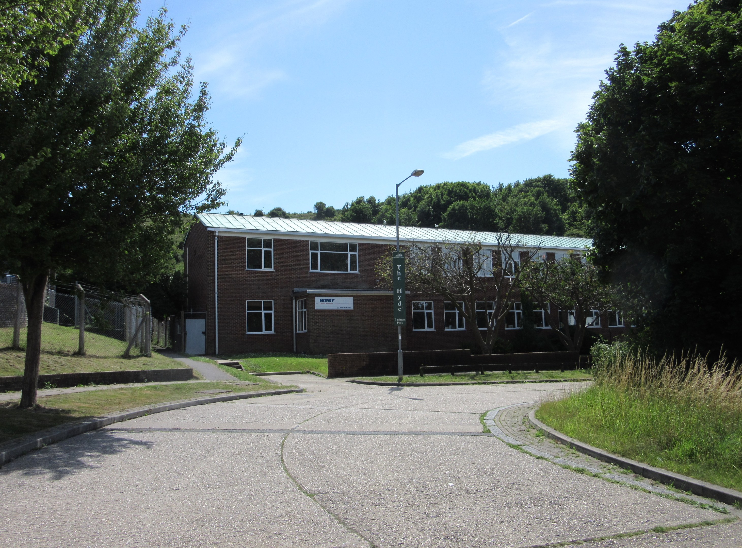Ein Schulgebäude mit Fenstern, ein Straßenmast, ein Schild, eine Baumgruppe, Gras, ein Zaun und ein bewölkter Himmel, umgeben von saftig grünem Gras in der Mitte eines Parks.