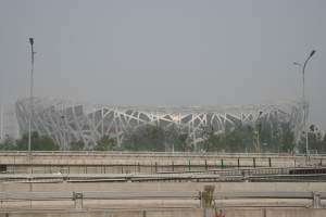 Olympiastadion in Peking mit Brücke, Geländern, Pfählen, Lichtern, Bäumen und einem Gebäude unter einem klaren blauen Himmel.