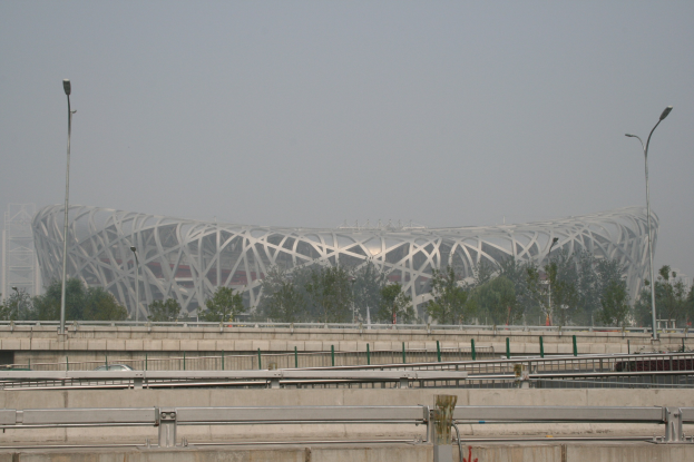 Olympiastadion in Peking mit Brücke, Geländern, Pfählen, Lichtern, Bäumen und einem Gebäude unter einem klaren blauen Himmel.