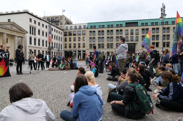 Eine Gruppe von Menschen, die auf dem Boden vor einer Menge sitzen, die Fahnen und Transparente schwenkt, mit einer Person, die in ein Mikrofon spricht, einer Statue und Gebäuden im Hintergrund während einer Anti-Schwulen-Demo in Berlin, Deutschland.