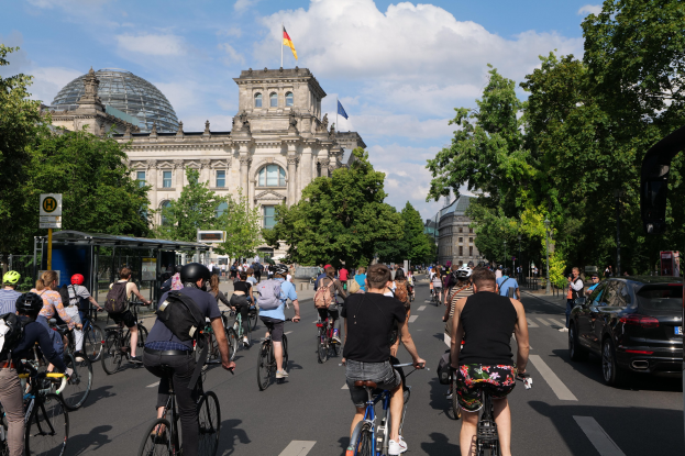 Gruppe von Menschen, die auf Fahrrädern eine von Bäumen gesäumte Straße in Berlin, Deutschland, entlangfahren, mit einer Bushaltestelle auf der rechten Seite und einer Fahne, die auf einem Gebäude weht, unter einem bewölkten Himmel.