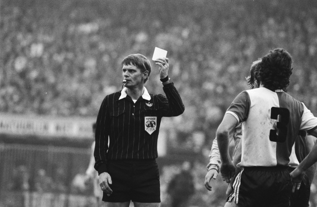 Ein Schiedsrichter in Schwarz-Weiß-Haltung hält eine Rote Karte vor einer Menge von Menschen in Fußballuniformen, mit einem Zaun im Hintergrund.