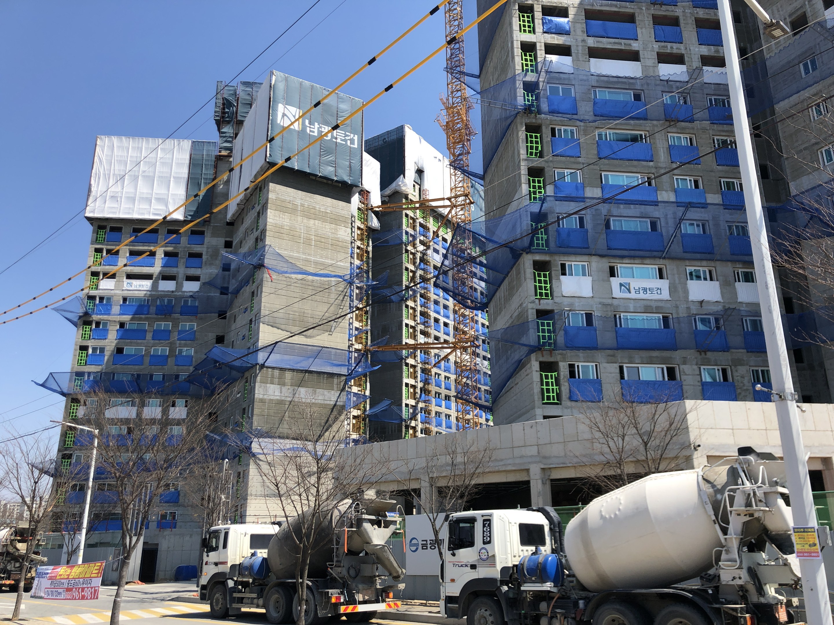Baustelle mit einem Betonmischfahrzeug, das vor einem hohen Gebäude unter Bauarbeiten geparkt ist, Fahrzeugen auf der Straße, Bäumen, Strommasten, Holzplanken, Kabeln im Vordergrund und Himmel im Hintergrund.