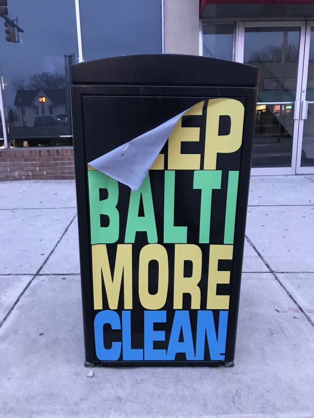 Ein schwarzer Müllcontainer mit "Keep Baltimore More Clean" darauf, vor einem Gebäude mit Glastüren auf dem Gehweg platziert.