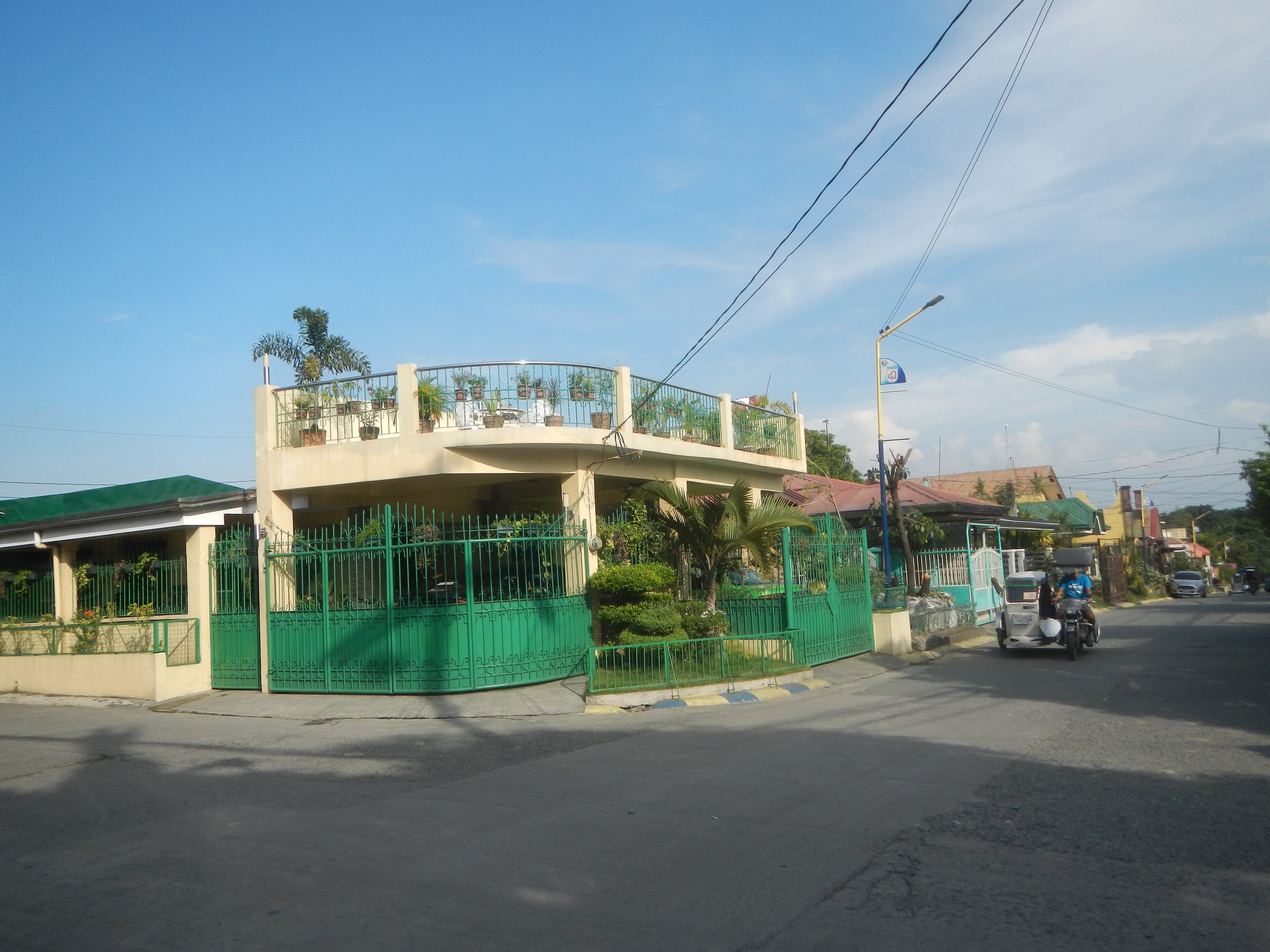 Straße mit Fahrzeugen, ein grünes Gebäude mit einem grünen Zaun, Geländern, Pflanzen, Bäumen, Pfählen, Lampen, Drähten und einer Flagge bei einem bewölkten Himmel.