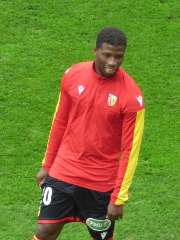Ein Mann in rotem Shirt und schwarzen Shorts auf einem Fußballfeld, der lächelt und eine Flügelposition einnimmt.