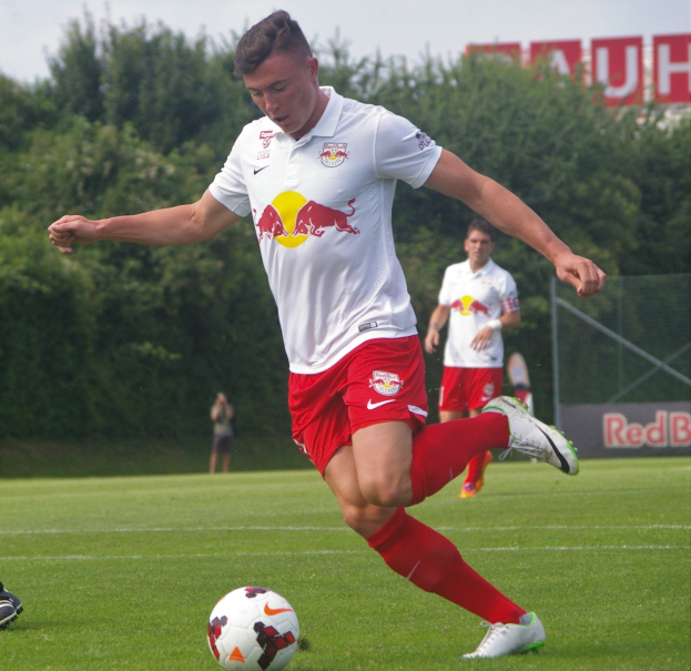 Ein professioneller Fussballspieler kickt einen Ball auf einem grünen Feld mit Bäumen und einem klaren blauen Himmel im Hintergrund, in der Nähe einer Tafel, auf der «RB Leipzig» steht.