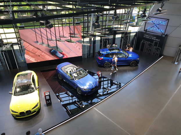Eine Gruppe von Menschen steht im BMW Museum in München, Deutschland, mit Autos auf der linken Seite und einem Schild mit der Aufschrift "BMW Museum München" auf der rechten Seite, mit Blick auf eine Außenansicht von Bäumen, Gebäuden und dem Himmel durch Glasfenster.
