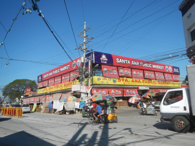 Eine belebte Straße mit einem Lastwagen, mehreren Fahrzeugen, Fußgängern, einem Strommast mit Überlandleitungen, Bäumen und einem klaren blauen Himmel.