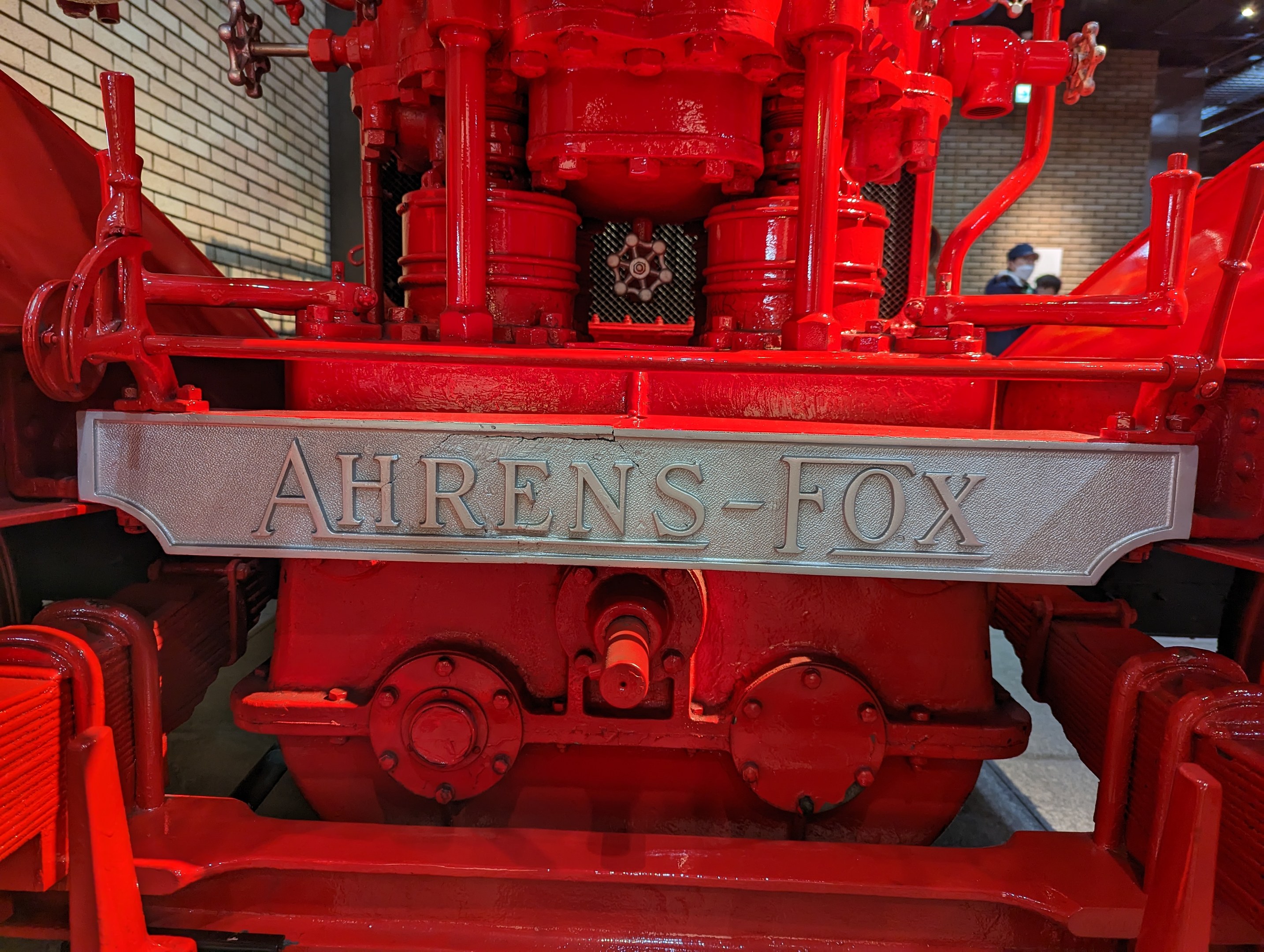 Nahaufnahme eines roten Feuerwehrautos in einem Museum, mit einer Wand links und einigen Menschen im Hintergrund unter Deckenbeleuchtung.