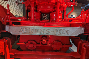 Nahaufnahme eines roten Feuerwehrautos in einem Museum, mit einer Wand links und einigen Menschen im Hintergrund unter Deckenbeleuchtung.