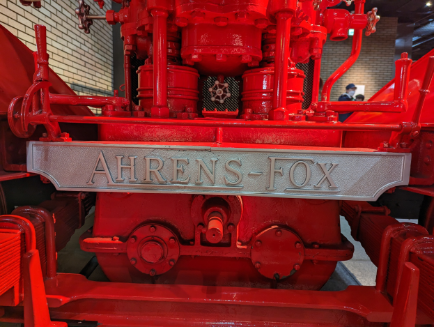 Nahaufnahme eines roten Feuerwehrautos in einem Museum, mit einer Wand links und einigen Menschen im Hintergrund unter Deckenbeleuchtung.