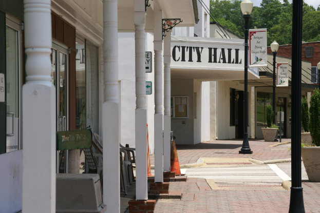 Ein Rathaus umgeben von Gebäuden, Straßenlaternen, Straßenschildern, Kraftfahrzeugen, Verkehrszeichen, Stühlen, Topfpflanzen, Bäumen und einem bewölkten Himmel.