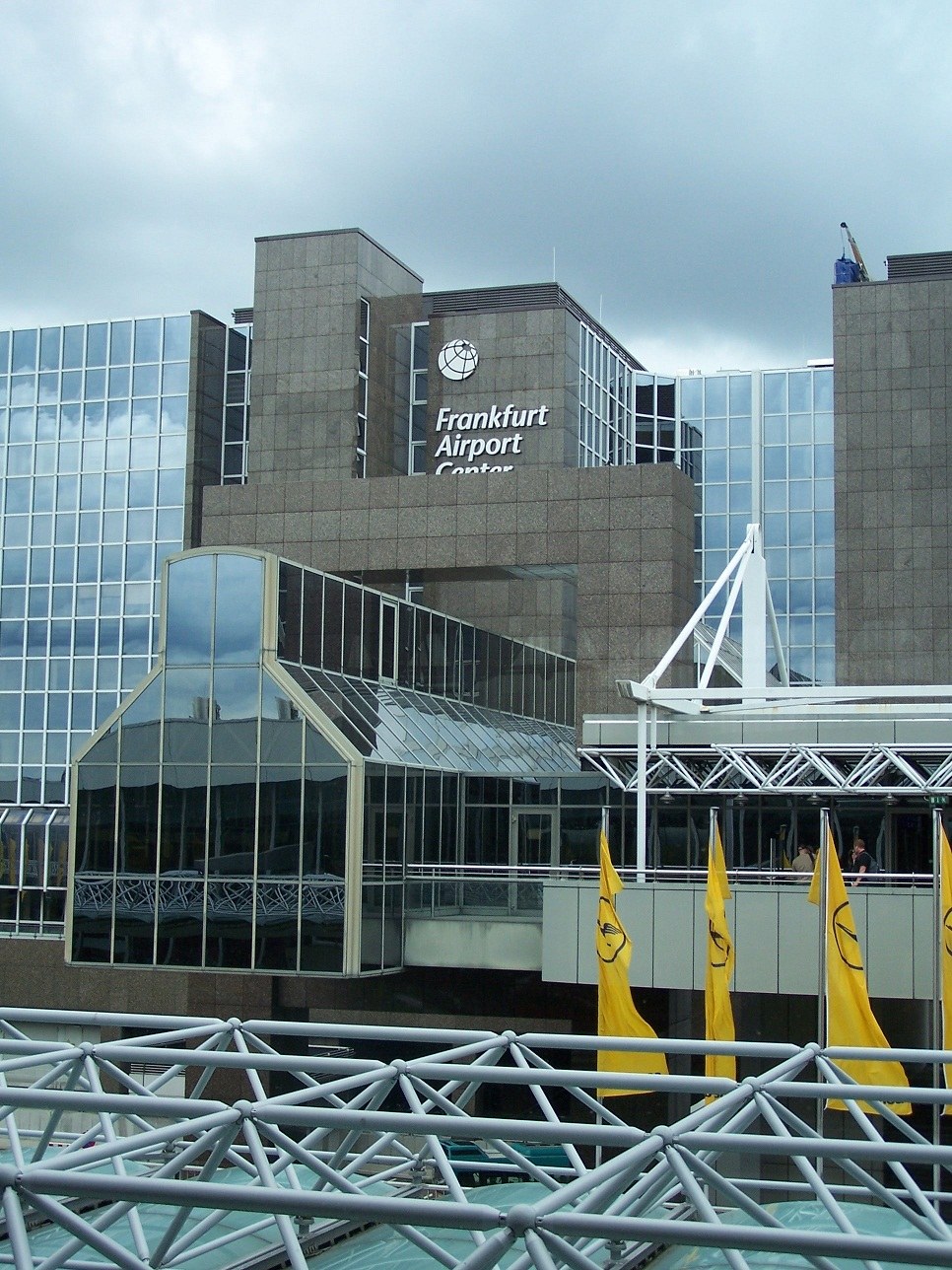 Frankfurt Airport mit Glaswänden, Text, gelben Flaggen und Eisenstangen unter einem bewölkten Himmel.