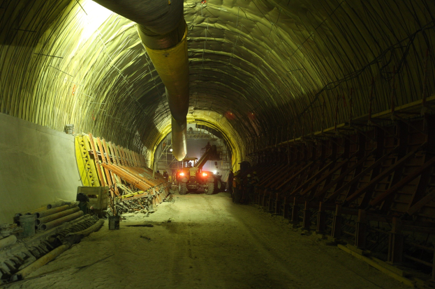 Großer Tunnel an einer Baustelle mit Fahrzeugen, verstreuten Materialien, einer Wand auf der linken Seite, Überkopfrohren und beleuchteter Hintergrundbeleuchtung.