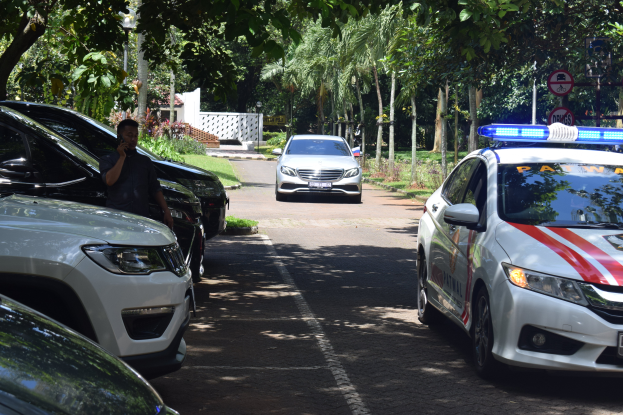 Ein Polizeiauto steht auf der Seite einer Straße neben anderen Fahrzeugen, mit einer Person auf der linken Seite, die ein Mobiltelefon hält, und einem Hintergrund mit Bäumen, Schildern, Pfählen, Pflanzen, Gras und einer Wand.