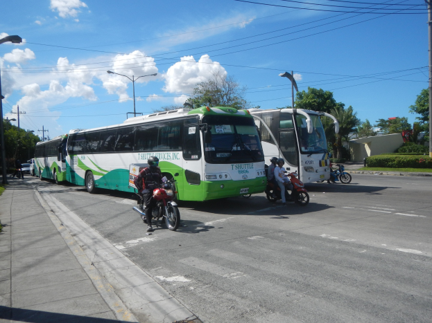 Ein grüner und weißer Shuttlebus neben einer Straße mit Motorradfahrern davor, einem grasbewachsenen Fußweg links und Gebäuden, Bäumen und Laternen im Hintergrund unter einem klaren blauen Himmel.