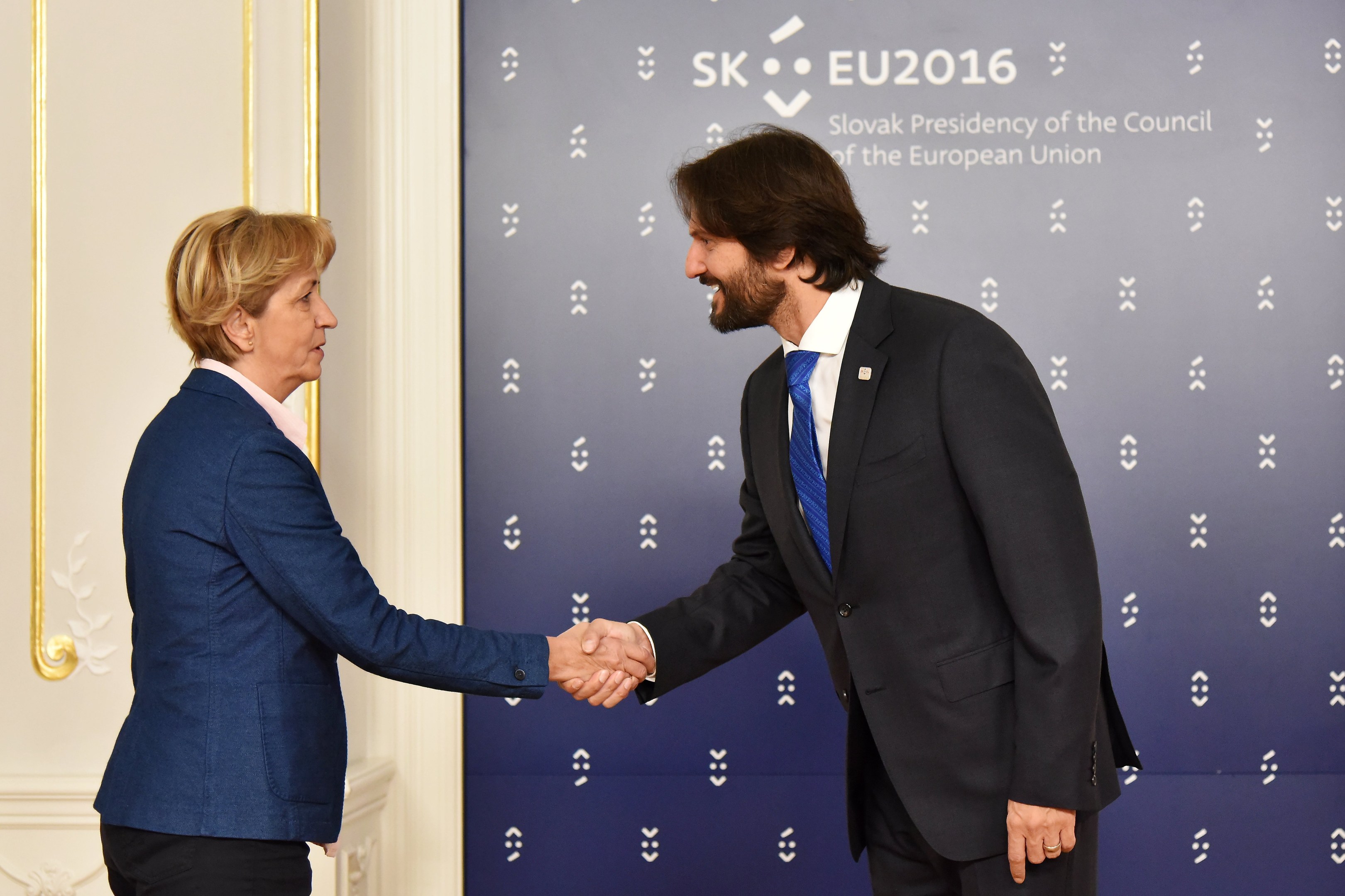 Bundeskanzlerin Angela Merkel und Bundespräsident Joachim Gauck geben sich die Hand vor einer Wand mit einem 'EU-Gipfel'-Schild und einer Tür im Hintergrund, beide lächeln.