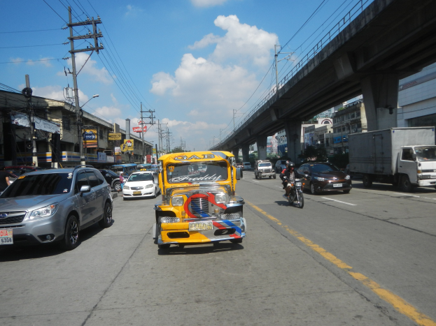 Ein gelbes Taxi fährt auf einer Straße mit hohen Gebäuden, Strommasten mit Drähten und einer Brücke im Hintergrund unter einem bewölkten Himmel mit einem Wasserzeichen auf dem Bild.
