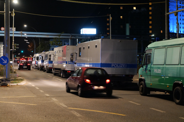 Lange Reihe von Polizeiwagen, die an einer Straße bei Nacht geparkt sind, mit sichtbarer Infrastruktur und Gebäuden im Hintergrund.