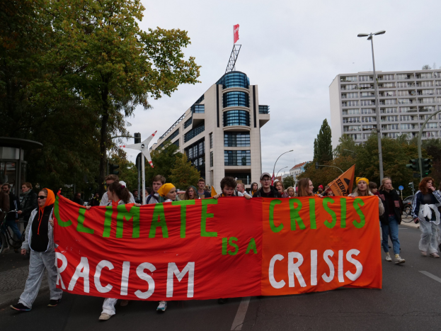 Eine Gruppe von Menschen geht die Straße entlang und hält ein Banner mit der Aufschrift "Klima-Krise ist eine Krise", mit Bäumen, Laternenmasten und Gebäuden an der Straße und dem Himmel im Hintergrund.
