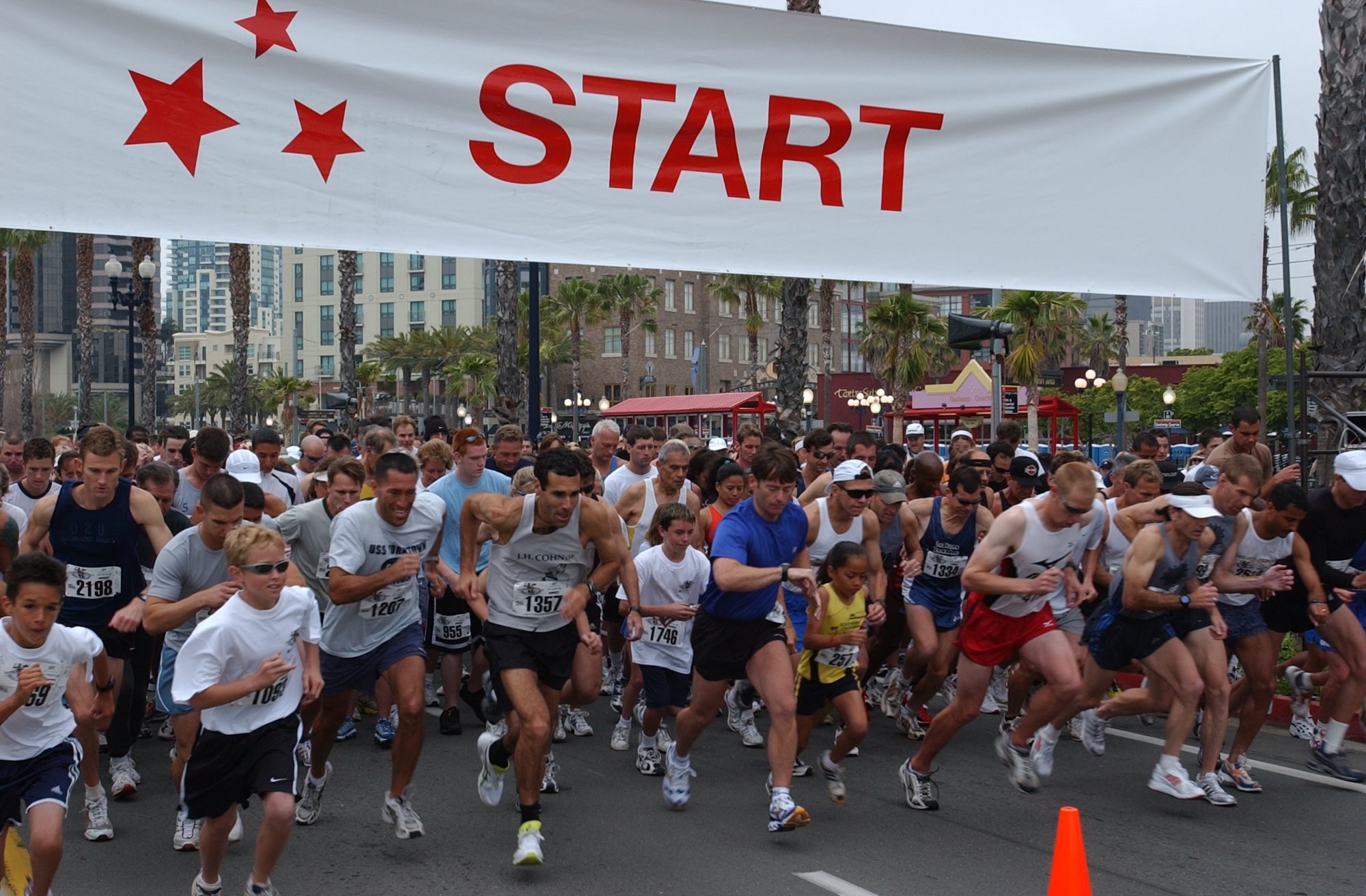 Gruppe von Menschen bei einem Marathon mit einem Verkehrskegel im Vordergrund und einem Banner im Hintergrund, umgeben von Bäumen, Laternenmasten, Gebäuden und einem klaren blauen Himmel.