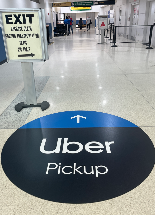 Ein Flughafen-Inneneinrichtung mit einem "Uber Pickup"-Schild auf dem Boden, Menschen im Hintergrund und verschiedene Gegenstände wie Bretter und Lampen.