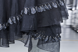 Nahaufnahme eines bodenlangen schwarzen Kleides auf dem Laufsteg mit hohem Kragen, langen Ärmeln, leichtem Stoff mit feiner Schimmer und kunstvoller Perlen- und Paillettenverzierung.