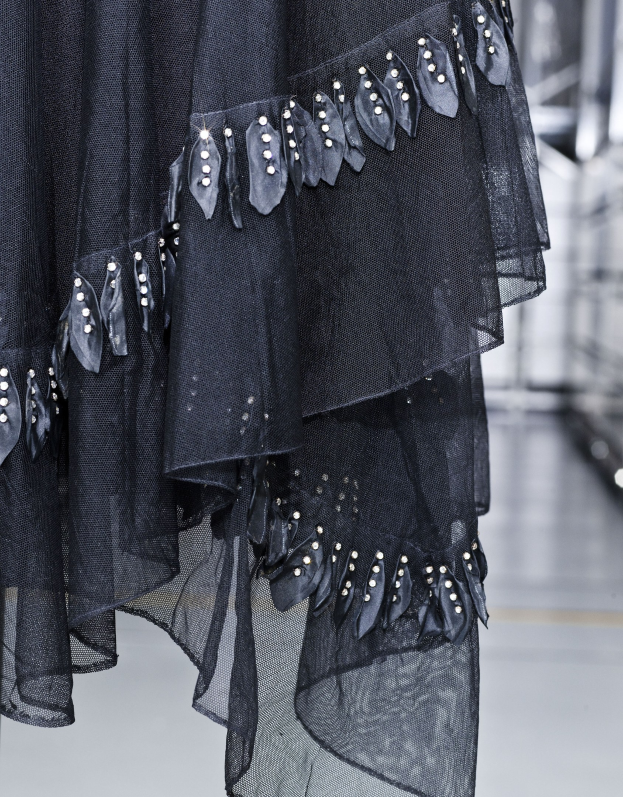 Nahaufnahme eines bodenlangen schwarzen Kleides auf dem Laufsteg mit hohem Kragen, langen Ärmeln, leichtem Stoff mit feiner Schimmer und kunstvoller Perlen- und Paillettenverzierung.