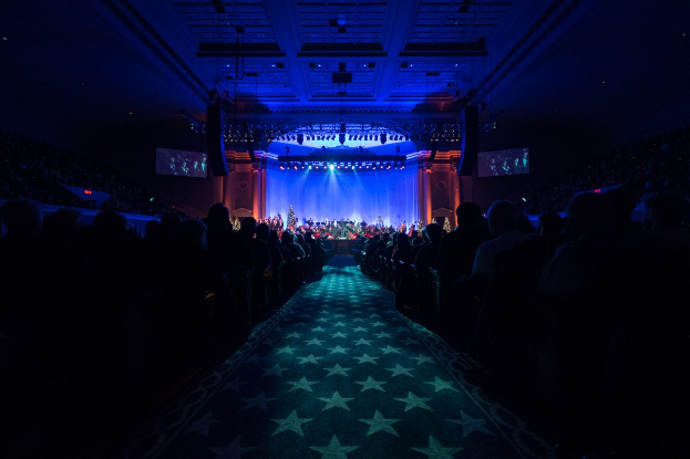 Ein großer Sitzungssaal mit sitzenden Gästen, eine Bühne mit Musikern und Deckenbeleuchtung und -fernseher, was auf ein 2019 Nationales Kongress-Konzerthaus hindeutet.