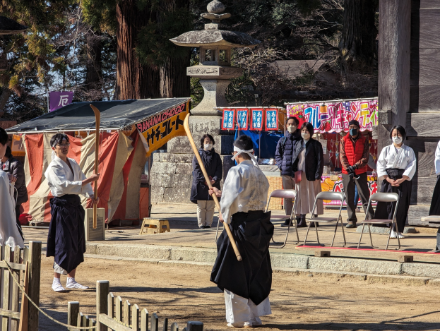 Eine Gruppe von Menschen in formeller Kleidung, einige mit Masken und Holzstöcken, versammelt sich im Freien vor einem traditionellen japanischen Gebäude während einer Zeremonie in Kyoto, mit Stühlen, Bannern, einem Zelt, Bäumen und einem klaren blauen Himmel.