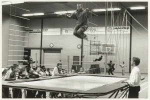 Eine Gruppe von Kindern sitzt um ein Trampolin in einer Turnhalle. In der Mitte springt eine Person in die Luft, während ein Junge rechts von ihnen steht. Unten befindet sich ein Tisch und im Hintergrund eine Wand mit einer Uhr und Tafeln mit Text.