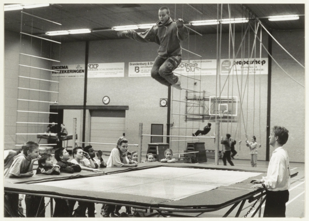 Eine Gruppe von Kindern sitzt um ein Trampolin in einer Turnhalle. In der Mitte springt eine Person in die Luft, während ein Junge rechts von ihnen steht. Unten befindet sich ein Tisch und im Hintergrund eine Wand mit einer Uhr und Tafeln mit Text.