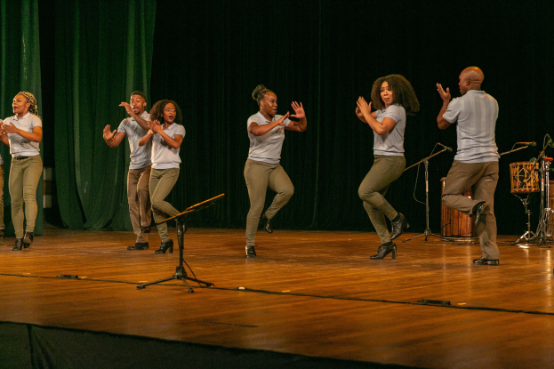 Eine Gruppe von Menschen auf einer Bühne, die applaudieren, mit Mikrofonen, Musikinstrumenten und einem Vorhang im Hintergrund, wahrscheinlich eine Tanzmannschaft, die ihren Erfolg anerkennt.