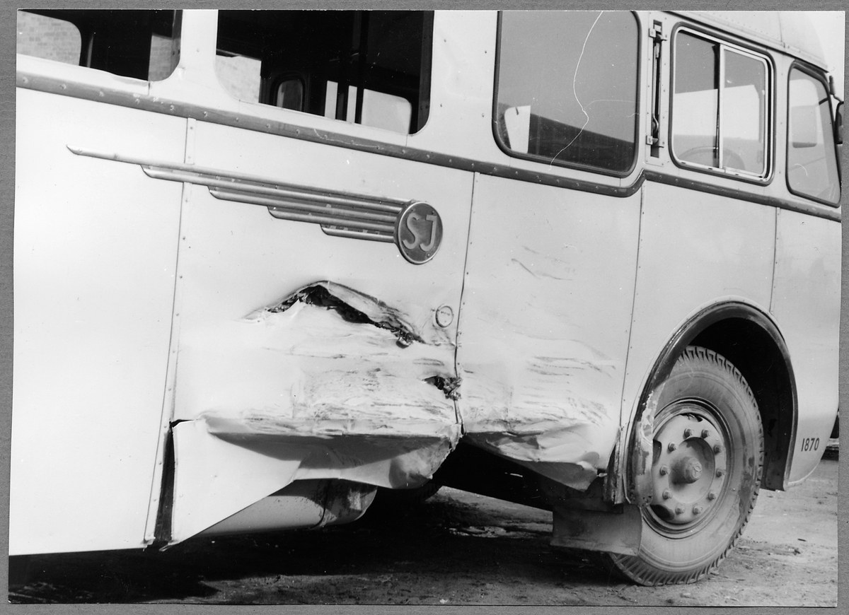 Schwarz-weißes Foto eines beschädigten Busses am Straßenrand mit eingedellter Front, zersplitterten Fenstern und offener Fahrertür, die den Innenraum zeigt.
