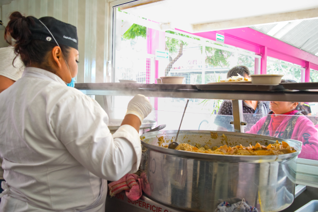 Eine Frau in Kochuniform, weißem Kleid, Handschuhen und schwarzer Mütze, hält einen Löffel und steht vor einer Stahlschüssel voller Essen, mit einer anderen Frau in blauem Kleid links von ihr und verschiedenen Küchenutensilien und Menschen im Hintergrund.